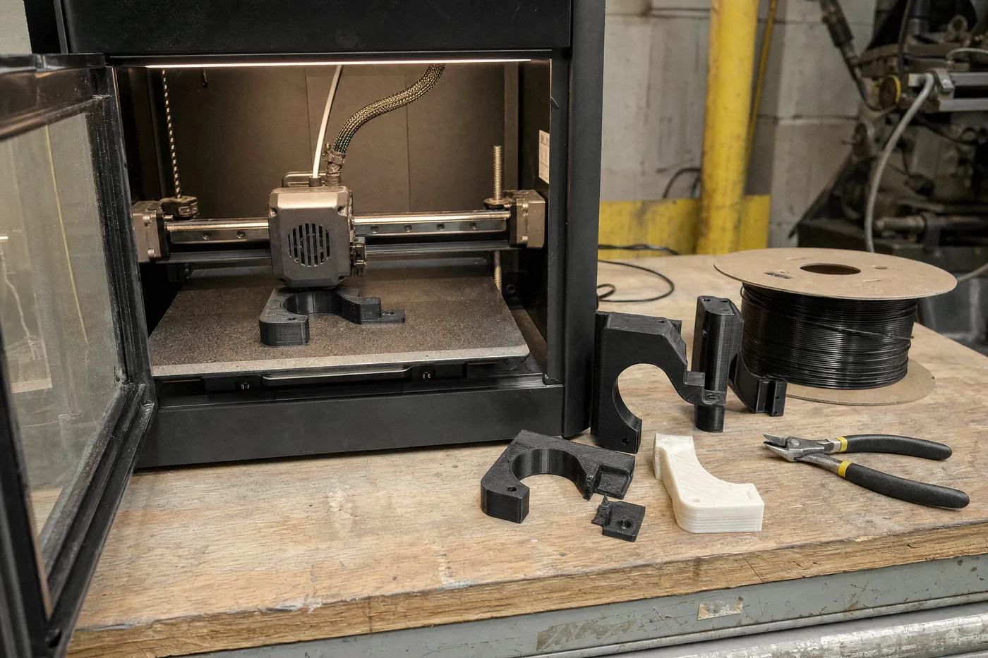 An enclosed 3D printer part-way through printing a black bracket. On the bench beside it are earlier versions of the same bracket, one snapped, and a spool of black filament.